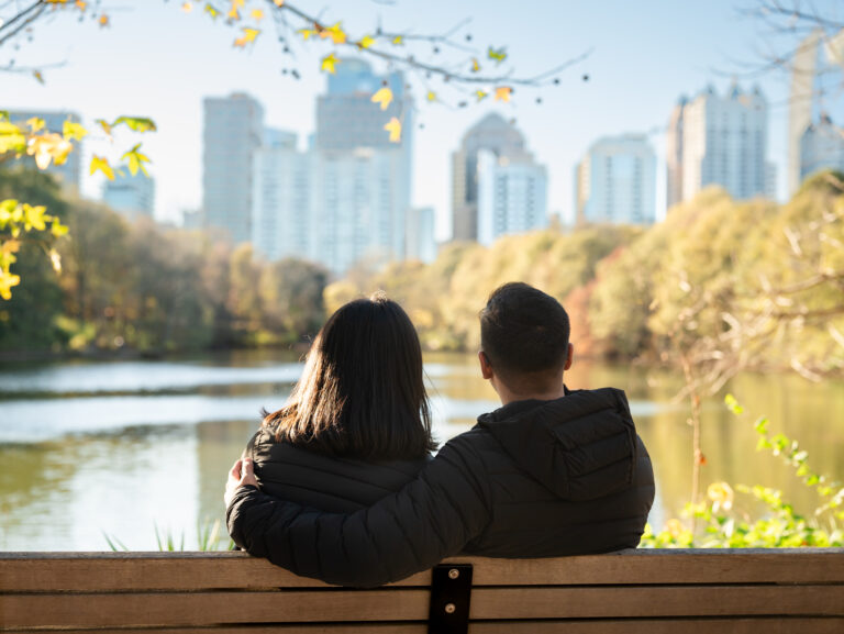 Couple sitting close on a bench overlooking the lake at Piedmont Park with the Atlanta skyline in the distance.