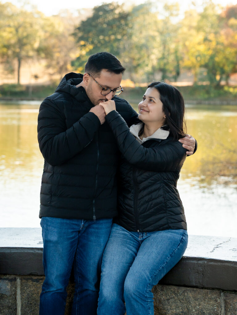 Engaged couple by the water at Piedmont Park in Atlanta as he kisses her hand and she smiles up at him on a stone ledge.