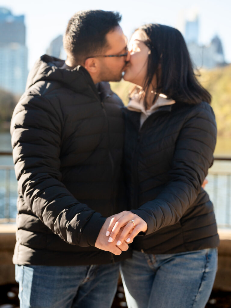 Newly engaged couple kissing by the lake with the Atlanta skyline softly blurred behind them at Piedmont Park.