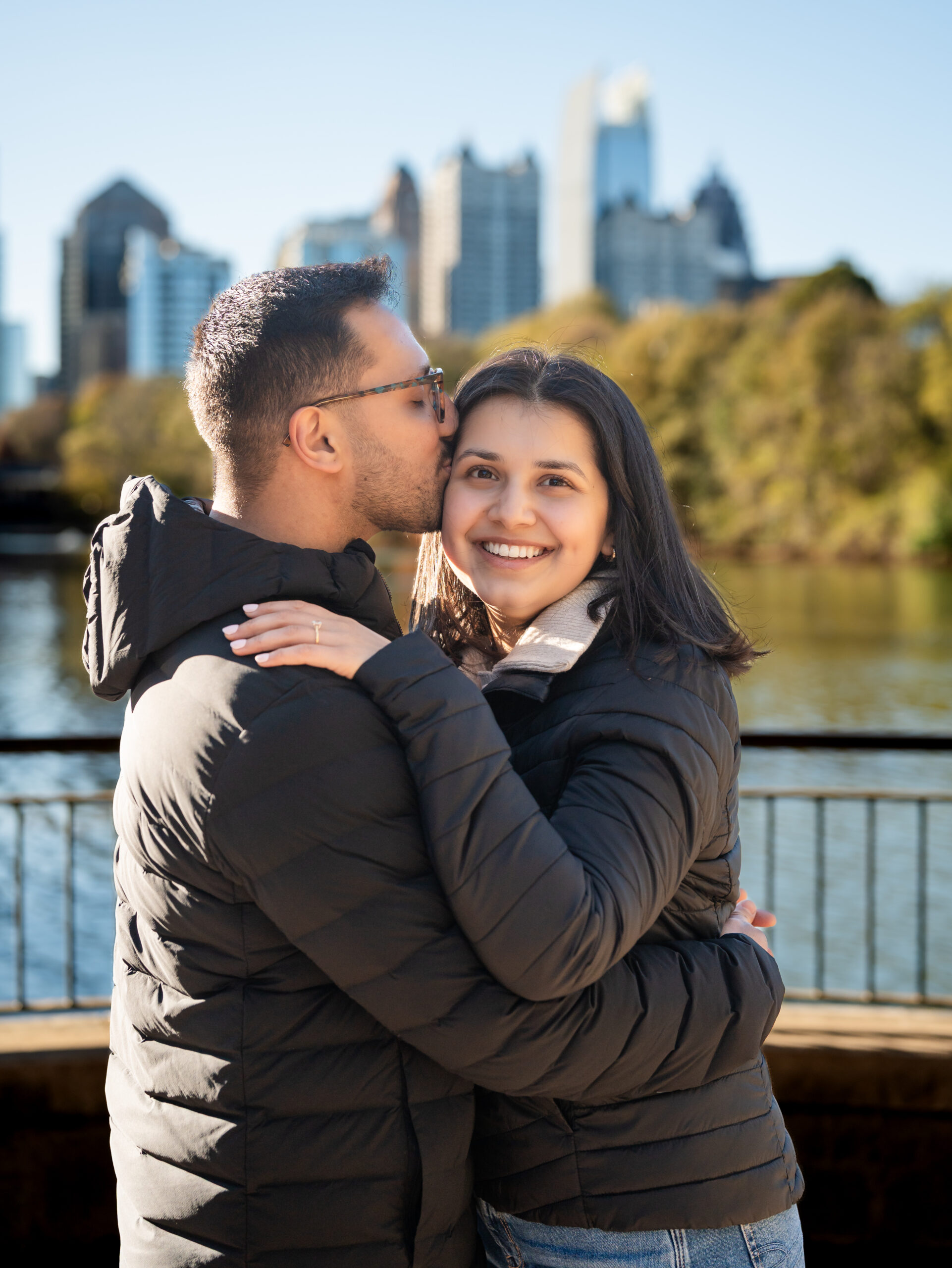 Newly engaged couple hugging by the lake at Piedmont Park with the Atlanta skyline in the background.