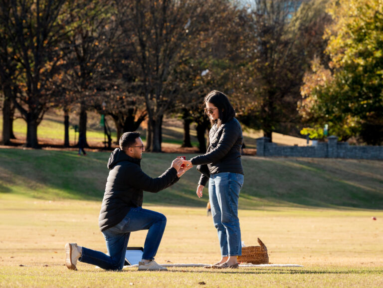 Surprise Piedmont Park proposal in The Meadow as he gets down on one knee near a cozy picnic setup.