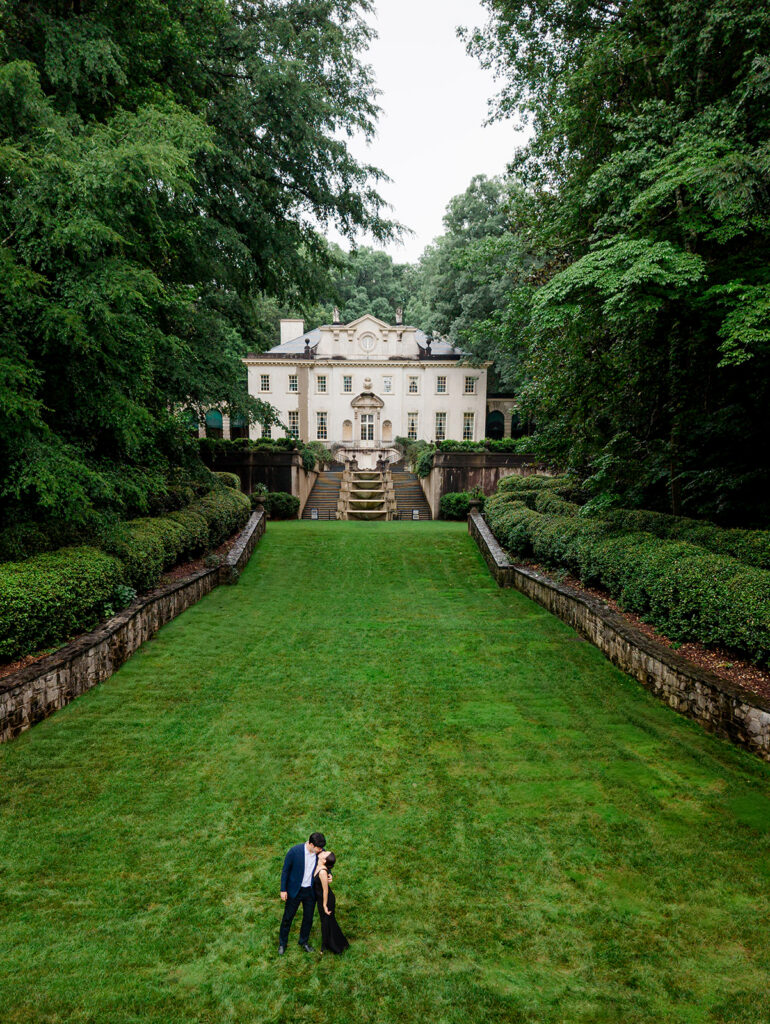 Aerial view of the couple kissing at the base of the long green lawn, hedges on both sides and the mansion at the top.