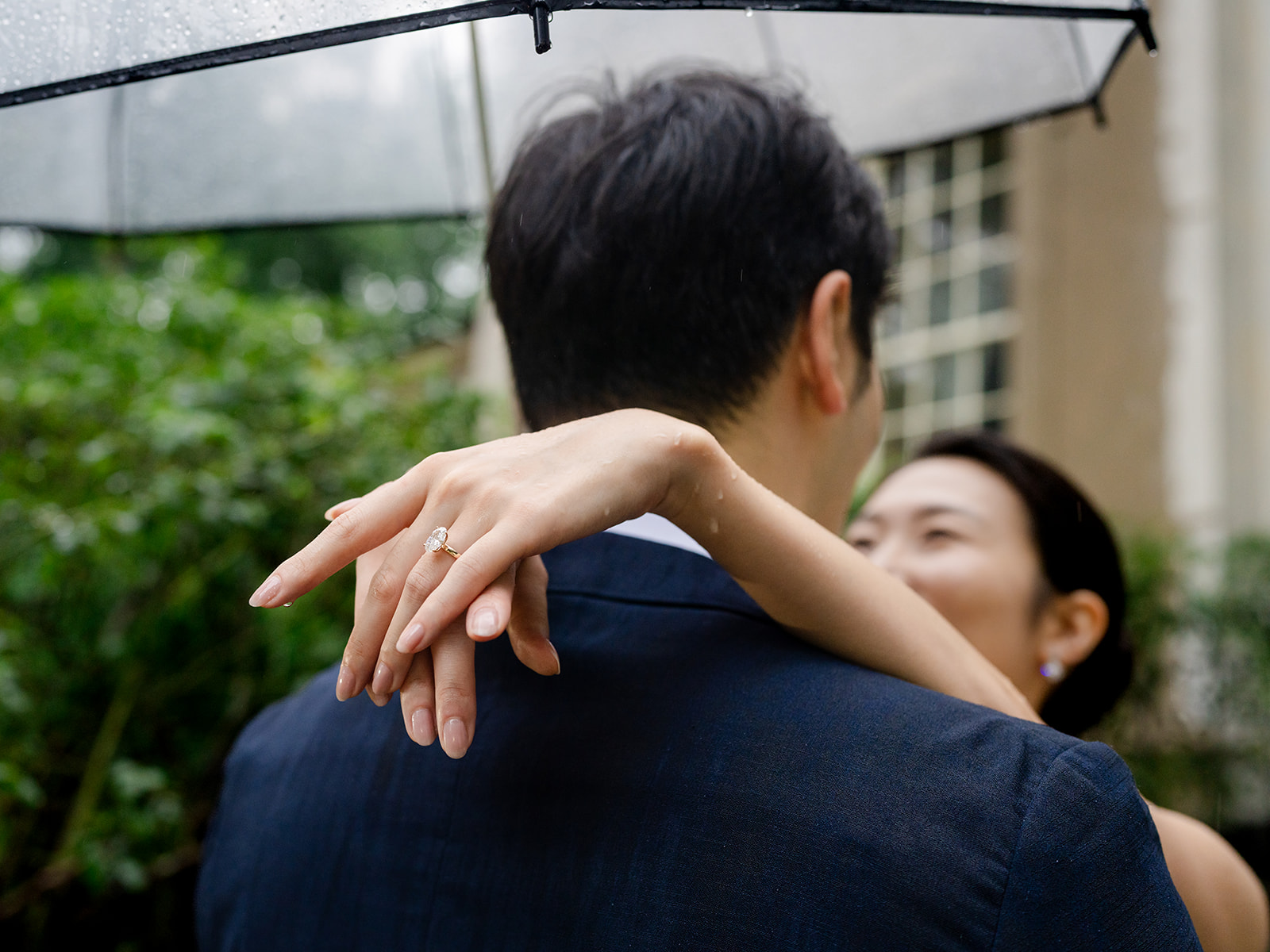 Oval diamond engagement ring on her hand with raindrops on her skin, her arm around his shoulder under a clear umbrella.