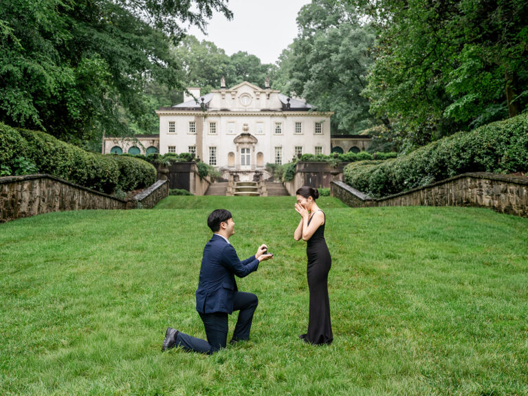 A Swan House Atlanta proposal on the lawn: he kneels with the open ring box as she lifts her hands to her face, the mansion behind them.